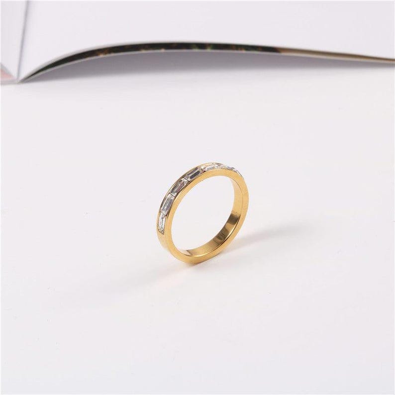May include: A gold-colored ring with rectangular, clear gemstones set around the band. The ring is on a white surface, with a blurred background.