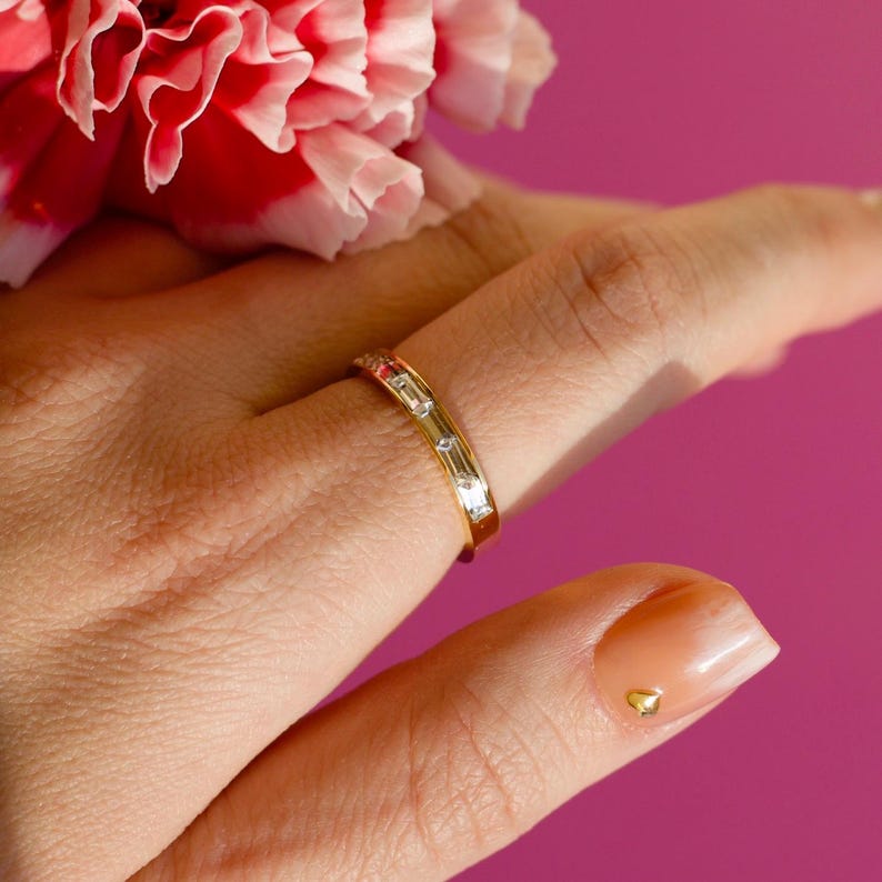 May include: A gold ring with rectangular and square-cut clear stones. The ring is on a finger, with a pink flower in the background. The nail has a gold heart-shaped decoration.
