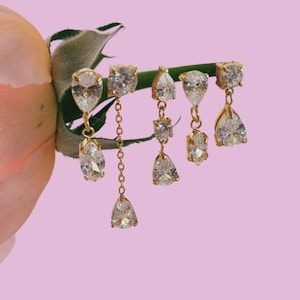 May include: Gold-toned earrings with clear, sparkling cubic zirconia stones. The earrings feature various shapes, including pear and square cuts, with some dangling from delicate chains. The jewelry is displayed against a soft pink background, with a rose stem in the background.