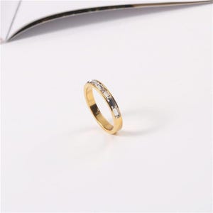May include: A gold-colored ring with rectangular, clear gemstones set in a row. The ring is on a white surface, with a curved, dark object in the background. The ring's band is a polished gold color.