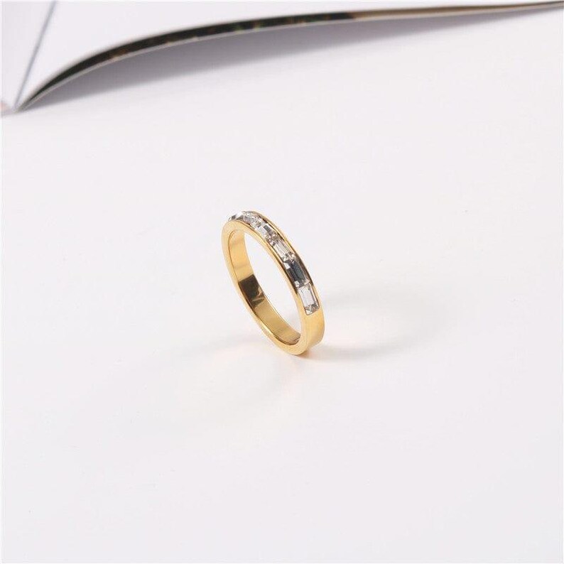 May include: A gold-colored ring with rectangular, clear gemstones set in a row. The ring is on a white surface, with a curved, dark object in the background. The ring's band is a polished gold color.