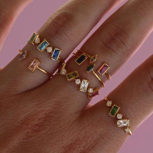 May include: Several gold rings adorned with colourful gemstones. The rings showcase a variety of gemstone shapes, including emerald-cut and round diamonds, in shades of blue, green, pink, and red. The rings are displayed against a pink backdrop.