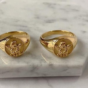Puede incluir: Dos anillos de sello dorados con un dise&ntilde;o de &aacute;ngel. Los anillos son redondos con un acabado pulido. Cada anillo presenta un relieve detallado de un querub&iacute;n con alas sobre una cara circular.