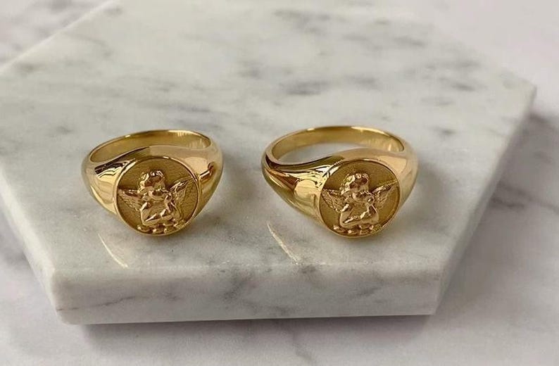 Puede incluir: Dos anillos de sello dorados con un dise&ntilde;o de &aacute;ngel. Los anillos son redondos con un acabado pulido. Cada anillo presenta un relieve detallado de un querub&iacute;n con alas sobre una cara circular.