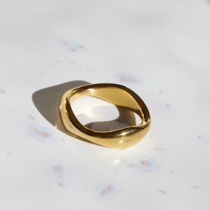 Peut inclure: Une bague dorée de forme irrégulière. La bague semble être faite d'un métal plein, avec une surface lisse et polie. Le design est moderne et minimaliste, avec une forme légèrement incurvée et rectangulaire. La bague est présentée sur une surface blanche.