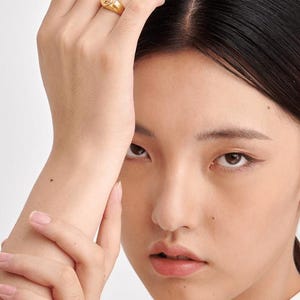 Puede incluir: Un anillo de sello dorado con un dise&ntilde;o detallado se muestra en un dedo. El anillo es el punto focal, sobre un fondo neutro. La imagen es un primer plano, que destaca los intrincados detalles del anillo y la textura de la piel.