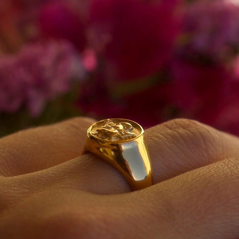 Puede incluir: Un anillo de sello dorado con un dise&ntilde;o en relieve detallado en la parte superior. El anillo se lleva en un dedo, con un fondo borroso de flores rosas. El anillo tiene una superficie pulida y reflectante.