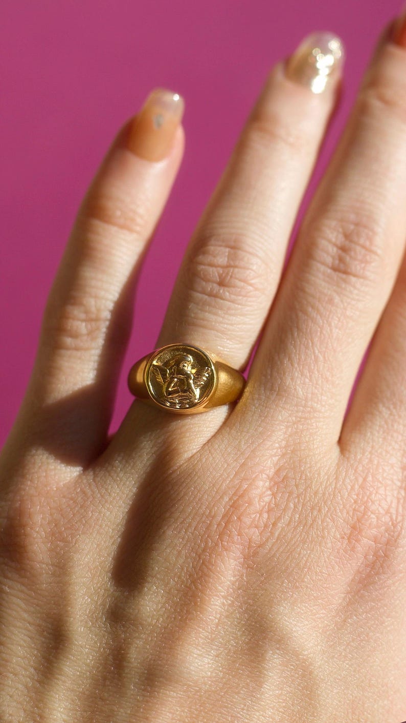 Puede incluir: Un anillo de sello dorado con un relieve detallado de un querub&iacute;n. El anillo se lleva en un dedo sobre un fondo rosa. El dise&ntilde;o del anillo es circular, con una banda lisa y pulida.