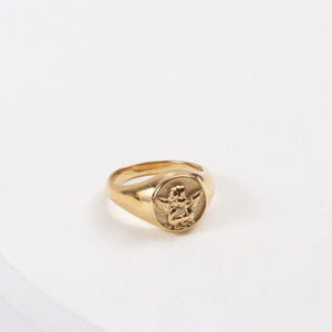Puede incluir: Anillo de sello dorado con un relieve detallado de un querub&iacute;n alado en la cara. El anillo tiene una banda lisa y pulida y una cara redondeada. El querub&iacute;n est&aacute; representado con alas extendidas y detalles intrincados.