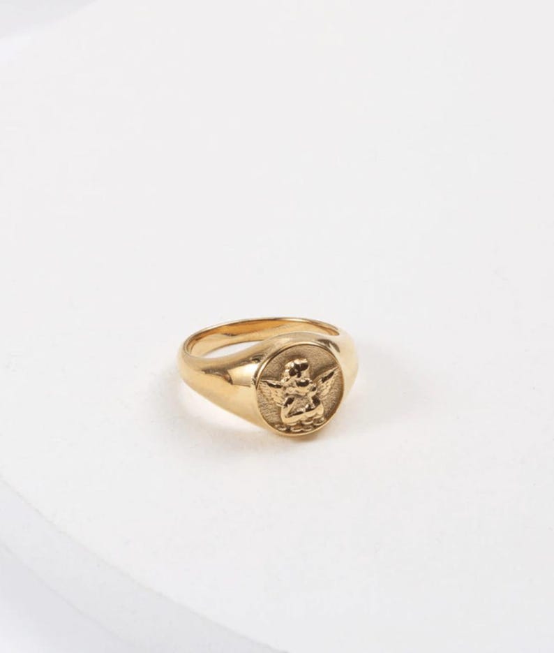Puede incluir: Anillo de sello dorado con un relieve detallado de un querub&iacute;n alado en la cara. El anillo tiene una banda lisa y pulida y una cara redondeada. El querub&iacute;n est&aacute; representado con alas extendidas y detalles intrincados.