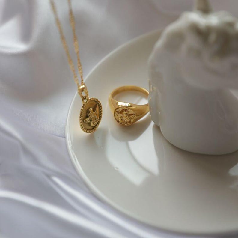 Puede incluir: Un collar dorado con un colgante de &aacute;ngel y un anillo dorado a juego, ambos con dise&ntilde;os de &aacute;ngeles. Las joyas se exhiben sobre un plato blanco y un fondo de sat&eacute;n blanco.