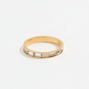 May include: A gold-colored ring with a band of rectangular, clear crystals. The ring is set against a white background, highlighting the sparkle of the crystals and the shine of the gold. The ring is a piece of jewelry.