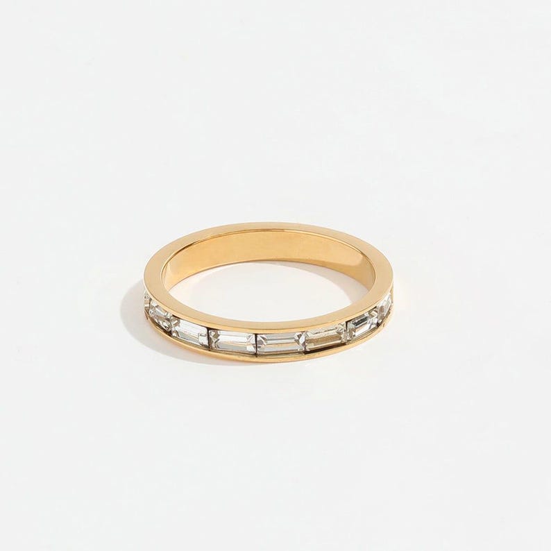 May include: A gold-colored ring with a band of rectangular, clear crystals. The ring is set against a white background, highlighting the sparkle of the crystals and the shine of the gold. The ring is a piece of jewelry.