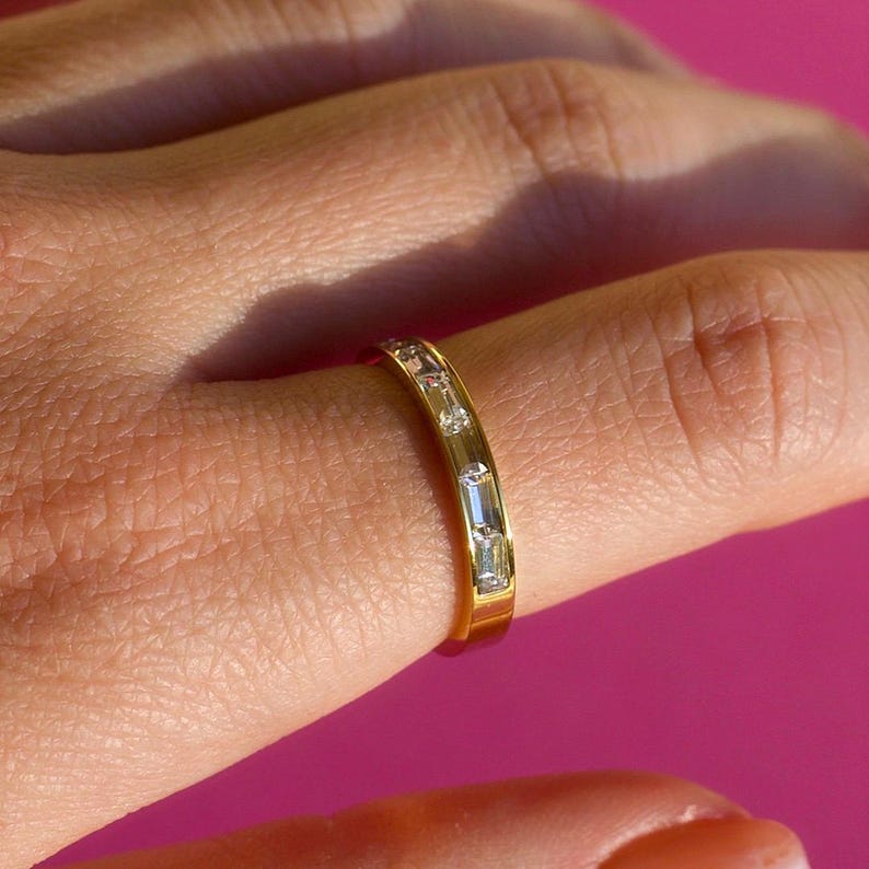 May include: A gold band ring with a series of clear, rectangular-cut gemstones. The ring is set on a finger against a bright pink background. The gemstones are evenly spaced around the band.