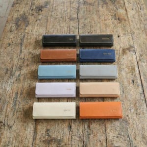 May include: A collection of rectangular, personalized leather wallets in various colors, including black, brown, blue, and orange. Each wallet features a name or initials embossed in gold lettering. The wallets are arranged on a rustic wooden surface.