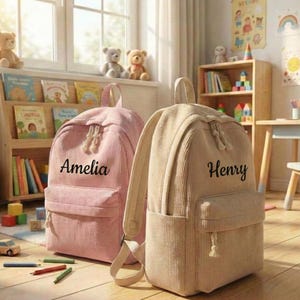 May include: Two corduroy backpacks, one pink with "Amelia" and one tan with "Henry" printed on them. Both have a front pocket and a top handle. The backpacks are in a child's room with toys and books.
