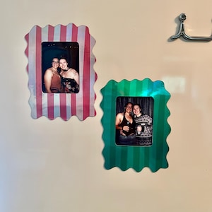 May include: Two decorative photo frames with scalloped edges are mounted on a wall. One frame is pink and white striped, the other is green and white striped. Each frame holds a photograph of people.