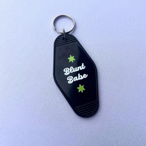 May include: Black motel-style keychain with the words "Blunt Babe" in white script, accented by two green star graphics. The keychain has a silver metal ring for attaching keys.