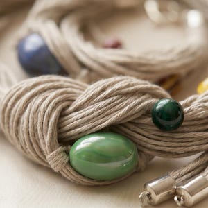 May include: Close-up of a beige rope necklace with a knot design. The necklace features a large, oval green bead, a small, round green bead, and a blue bead. Silver-colored metal clasps are visible at the end of the rope.