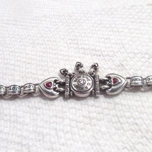 May include: A silver-toned metal chain bracelet featuring repeating links engraved with the words LOVE, JOY, and HOPE. The center piece is a circular charm with the text A HEART BEAT IS A LOVE BEAT, flanked by two heart-shaped charms with red accents.