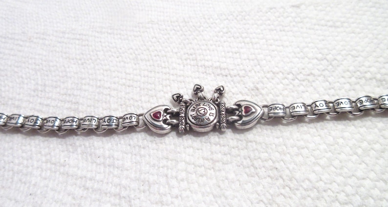 May include: A silver-toned metal chain bracelet featuring repeating links engraved with the words LOVE, JOY, and HOPE. The center piece is a circular charm with the text A HEART BEAT IS A LOVE BEAT, flanked by two heart-shaped charms with red accents.