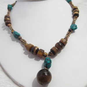 May include: A handcrafted necklace featuring a combination of brown tiger eye beads, turquoise stones, and gold-toned metal accents. The necklace has a pendant with a large, round tiger eye bead. The beads are strung together to create a unique and stylish accessory.