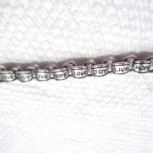May include: A silver-toned metal charm bracelet featuring a series of cylindrical beads, each engraved with the words LOVE, JOY, LIVE, or HOPE in black lettering. The bracelet consists of interconnected circular links and a heart-shaped charm.