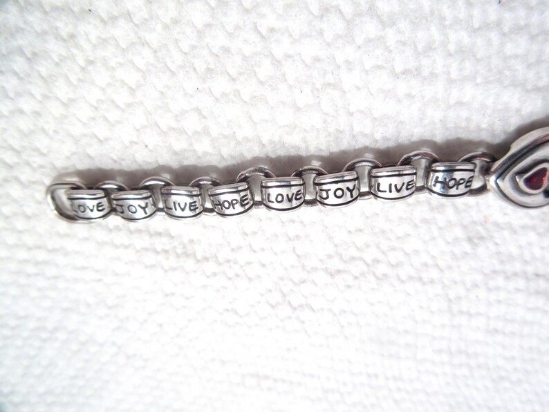 May include: A silver-toned metal charm bracelet featuring a series of cylindrical beads, each engraved with the words LOVE, JOY, LIVE, or HOPE in black lettering. The bracelet consists of interconnected circular links and a heart-shaped charm.