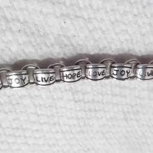 May include: A silver-toned chain bracelet featuring cylindrical metal beads. Each bead is engraved with the words LOVE, JOY, LIVE, or HOPE in black lettering. The bracelet consists of interconnected circular links holding the beads in a repeating sequence.