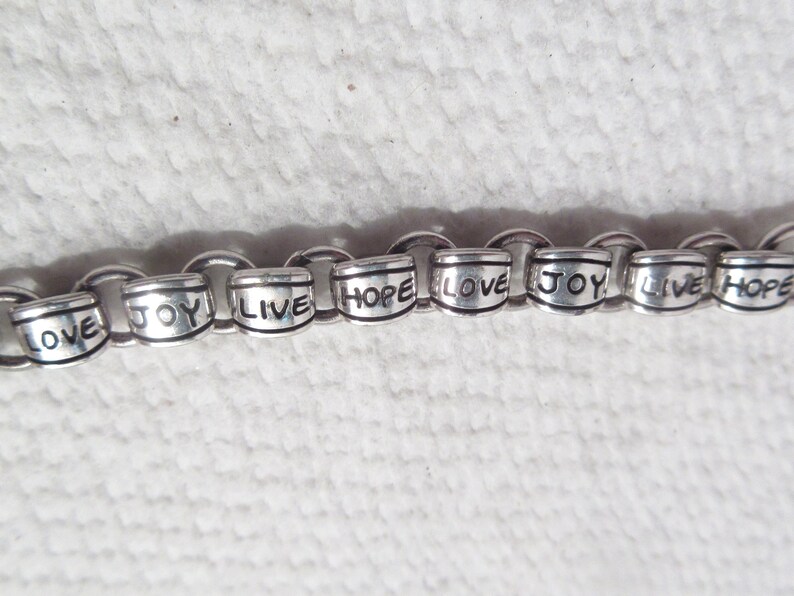 May include: A silver-toned chain bracelet featuring cylindrical metal beads. Each bead is engraved with the words LOVE, JOY, LIVE, or HOPE in black lettering. The bracelet consists of interconnected circular links holding the beads in a repeating sequence.