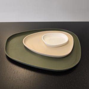 Könnte beinhalten: Ein Set aus drei ineinander stapelbaren Schalen mit unregelmäßigen, abgerundeten dreieckigen Formen. Die größte Schale ist olivgrün, die mittlere beige und die kleinste, flachste Schale ist weiß. Jede Schale hat eine glatte, matte Oberfläche.