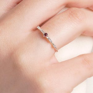 May include: A delicate silver ring featuring a small, round, deep red gemstone at the center, flanked by smaller, clear gemstones. The ring is set on a slender band, creating a minimalist and elegant design. The ring is shown on a person's finger.