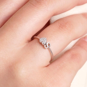 Puede incluir: Un anillo de plata con un diseño en forma de corazón incrustado con pequeñas piedras preciosas transparentes. El anillo también incluye una letra "S" estilizada en la banda. El anillo se muestra en un dedo.