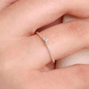 Puede incluir: Un anillo fino de metal plateado pulido con un delicado diseño en forma de V en el centro. La punta del diseño está acentuada con tres pequeñas piedras claras de talla redonda dispuestas en grupo, creando un aspecto sutil y refinado.