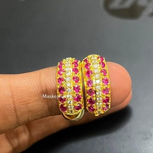 Könnte beinhalten: Ein Paar goldene Creolen mit einer mittleren Reihe aus klaren Steinen, flankiert von zwei Reihen leuchtend pinker runder Steine. Der Text Maskejewels ist auf der Oberfläche des Goldmetalls zwischen den beiden Ohrringen zu sehen.
