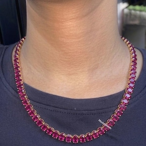 May include: A gold-toned tennis necklace featuring a continuous row of oval-cut, vibrant magenta-pink gemstones. The necklace rests against the neck, showcasing a uniform setting where each stone is secured in a metal frame, creating a linked chain.