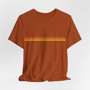 May include: A rust-colored t-shirt featuring a horizontal stripe design across the chest. The stripes are in shades of orange and yellow. The shirt has short sleeves and a crew neck.
