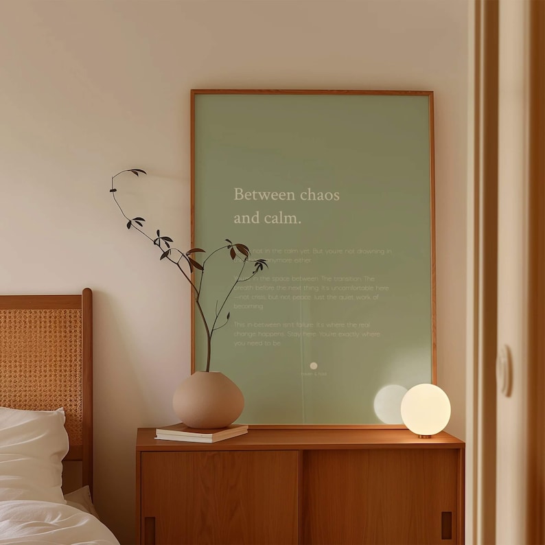 Może przedstawiać: Oprawiony pionowy plakat ścienny z szałwiowym tłem i białym napisem "Between chaos and calm." Wydruk znajduje się w cienkiej drewnianej ramie, ustawiony na drewnianej szafce obok okrągłego wazonu i małej lampy w kształcie kuli.