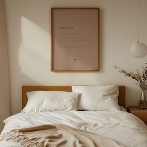 Op de afbeelding: Een ingelijste wandkunstprint met een oudroze achtergrond met het woord Sanctuary in vetgedrukte zwarte tekst, gevolgd door een definitie in woordenboekstijl. De print zit in een dunne houten lijst boven een bed met wit beddengoed.