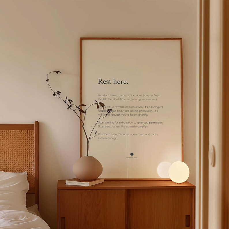 以下が含まれることがあります： 「Rest here.」というテキストと休息に関する段落が書かれた縦型の額入りウォールアート。木製のキャビネットの上に置かれ、細い枝が生けられたマットなベージュの花瓶と、小さく光る球形の白いテーブルランプが隣に配置されています。
