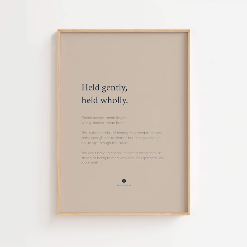 May include: A rectangular wall art print in a light wood frame featuring a beige background with dark blue text. The text reads: Held gently, held wholly. Gentle doesn't mean fragile. Wholly doesn't mean fixed. This is the paradox of healing.
