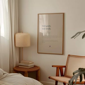May include: A rectangular wall art print in a thin wooden frame featuring the text Held gently, held wholly. The print is displayed on a light-colored wall above a small round wooden side table with a stack of books and a textured lamp.