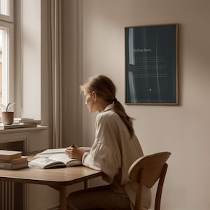 May include: A rectangular wall art print in a thin wooden frame featuring a dark navy blue background with the white text "Soften here." and smaller body text below. The print is displayed on a plain wall above a wooden desk and chair.