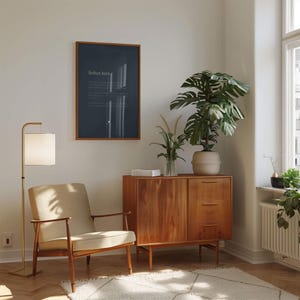 May include: A framed dark blue wall art piece with the text 'Soften here' displayed above a wooden sideboard, a beige upholstered armchair with wooden arms, a floor lamp with a white shade, and a potted monstera plant on the sideboard.