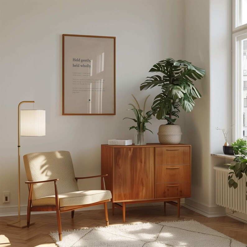 May include: A beige upholstered armchair with wooden armrests, a wooden cabinet with three drawers and a door, a floor lamp with a white shade, and a framed wall print with the text Held gently, held wholly, alongside a potted monstera plant.