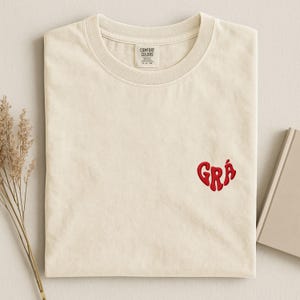 Könnte beinhalten: Cremefarbenes T-Shirt mit einem roten, herzförmigen, gestickten Design mit dem Wort "Grá". Das Shirt besteht aus einem weichen Material und hat einen klassischen Rundhalsausschnitt. Das Etikett lautet "Comfort Colors".