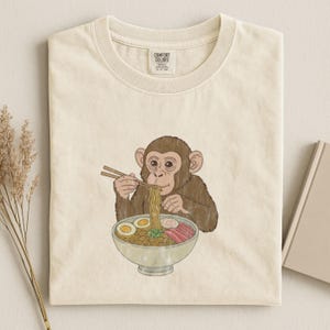 以下が含まれることがあります： 茶色の猿が箸を使ってラーメンを丼から食べているイラストが描かれたクリーム色のTシャツ。ラーメンには麺、卵、肉が含まれています。シャツにはComfort Colorsのタグが付いています。