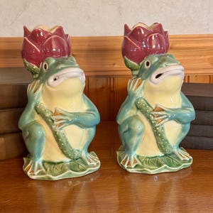 May include: A pair of ceramic frog figurines, each featuring a glossy finish with a pale yellow belly, teal limbs, and a dark red lotus flower resting on its head. The frogs are depicted in a seated position, holding a textured green stem.