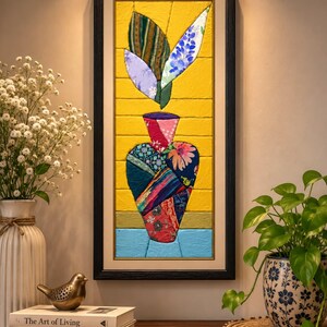 May include: A rectangular framed wall art piece featuring a fabric collage of a vase filled with leaves. The vase is constructed from various patterned fabric scraps in shades of red, blue, and pink, set against a bright yellow background.