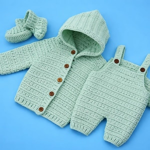 Puede incluir: Un conjunto de bebé de ganchillo verde claro, que incluye una chaqueta con capucha con botones de madera, un peto y patucos. El conjunto se muestra sobre un fondo azul. El punto de ganchillo crea una apariencia texturizada.